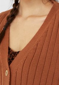 Cardigan in roestkleur met een geribbelde textuur, V-hals ontwerp en knoopsluiting. Daaronder is een donkergekleurd patroon te zien.