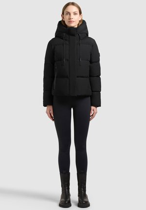 Jeune femme debout portant une doudoune noire avec capuche, des leggings noirs et des bottes noires sur un fond gris clair uni.