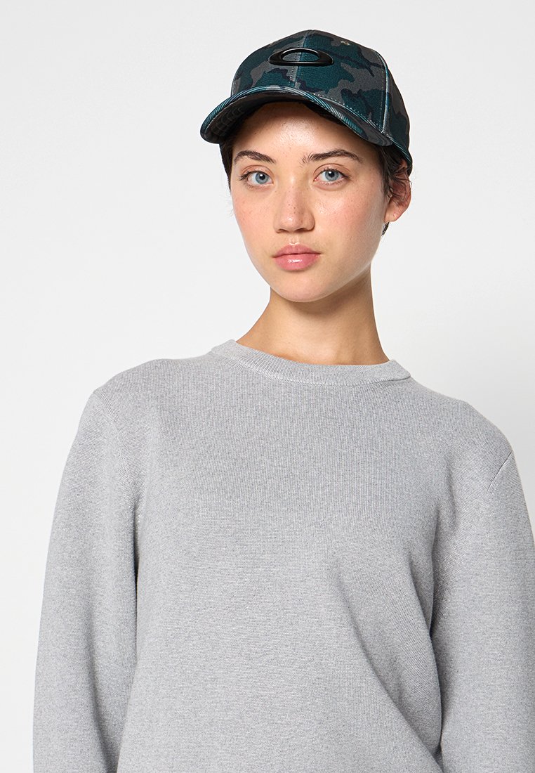 Gorra con estampado de camuflaje y visera curvada, que presenta un acento de logo metálico. Lleva puesta un suéter de cuello redondo texturizado en gris claro.