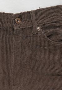 Close-up van de tailleband van donkerbruine corduroypantalon met een metalen knoop en klinknagel bij een zijzak, gedragen met een witte top.