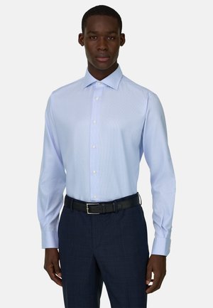 Mand iført lyseblå skjorte med knapper, stoppet ned i mørkeblå bukser med sort bælte, stående mod en ensfarvet baggrund.