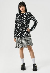 Femeie în picioare, purtând o rochie lungă cu mâneci lungi alb-negru, cu imprimeu cu inimă în partea superioară și fustă cu imprimeu houndstooth, șosete albe și pantofi negri cu cataramă aurie.