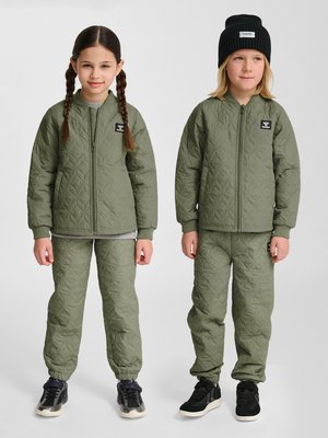 Twee kinderen staan naast elkaar in bijpassende olijfgroene doorgestikte jacks en broeken; het ene kind heeft vlechten, het andere draagt een zwarte muts.