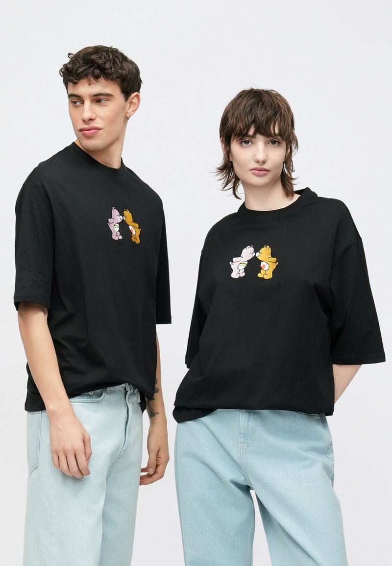 Μαύρα κοντομάνικα μπλουζάκια με καρτούν γραφικά αρκούδων σε ροζ και κίτρινο. Κανονική εφαρμογή, συνδυασμένα με ανοιχτόχρωμο μπλε τζιν.