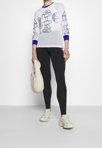 Bílý síťovaný top s dlouhým rukávem a modrými akcenty, zdobený výrazným nápisem "Adidas". Kombinován s černými legínami a béžovými sportovními botami. Drží měkkou tašku.