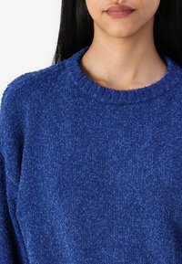 Pull bleu texturé avec un col rond. Le tissu semble doux et moelleux, avec une coupe légèrement ample et des détails côtelés au niveau du col.