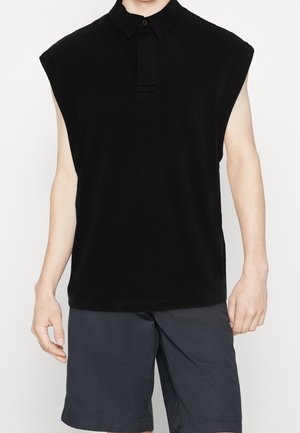 Personne portant un polo noir sans manches et un short gris foncé, se tenant avec les bras détendus le long du corps.