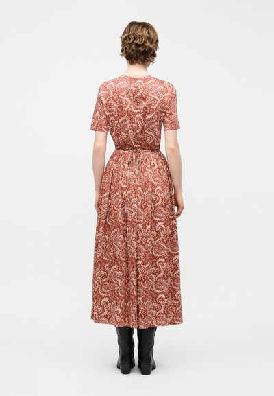 Γυναίκα με κοντά, σγουρά μαλλιά, φοράει μεσοφόρεμα με μοτίβο λουλουδιών σε σκουρόχρωμο πορτοκαλί και μαύρες μποτάκι αστραγάλου, στρέφεται με την πλάτη.