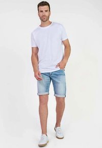 Camiseta blanca de algodón combinada con pantalones cortos de mezclilla azul claro, doblados en el dobladillo. Completo con zapatillas blancas y una pose casual de pie.