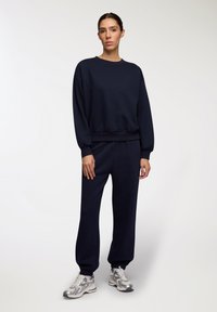 Granatowy zestaw bluzy i spodni dresowych z miękkiego materiału, z okrągłym dekoltem, luźnym krojem i elastycznymi mankietami. W zestawie z srebrno-białymi trampkami.