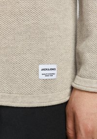 Beżowy sweter dzianinowy z teksturowanym wzorem, z ribbami u rękawów i białą metką z napisem "JACK & JONES" informującą o "Jakości Podstawowej od 1990 roku."