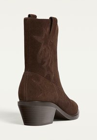 Bottine en suede marron avec un talon effilé, motif floral en relief et languettes latérales pour un enfilage facile. Texture lisse avec une semelle robuste.