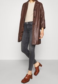 Kobieta ubrana w brązowy skórzany płaszcz, beżowy strukturalny top, czarne obcisłe dżinsy oraz brązowe klapki na obcasie stoi na tle jednolitego tła.