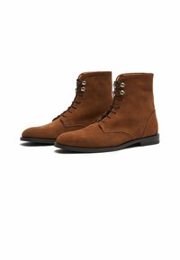 Botas de tobillo de ante marrón con una textura suave, diseño con cordones y ojales de metal, que presentan una puntera redondeada y una suela plana de goma.