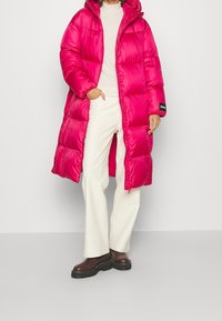 Abrigo largo acolchado en un vibrante color rosa, con capucha, cierre de cremallera y bolsillos laterales. Combinado con pantalones beige y zapatos plataforma marrón oscuro.