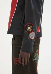 Sort læderjakke med røde detaljer, prydet med broderede lapper af et skull og flammer på ærmet. Tekstureret stof og tætsiddende design.