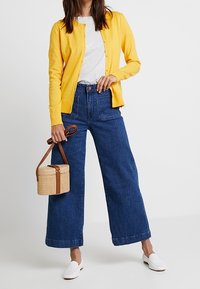 Gul stickad kofta, ljusgrå t-shirt, vida mörkdenimjeans, vita slip-on skor och en liten korgväska med bruna accenter.