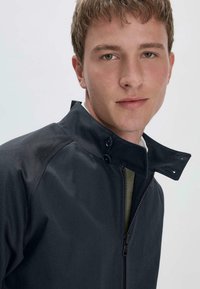 Zwarte jas met een staande kraag, voorzien van een rits en drukknoopsluiting, gemaakt van gladde stof met een contrasterende binnenvoering.