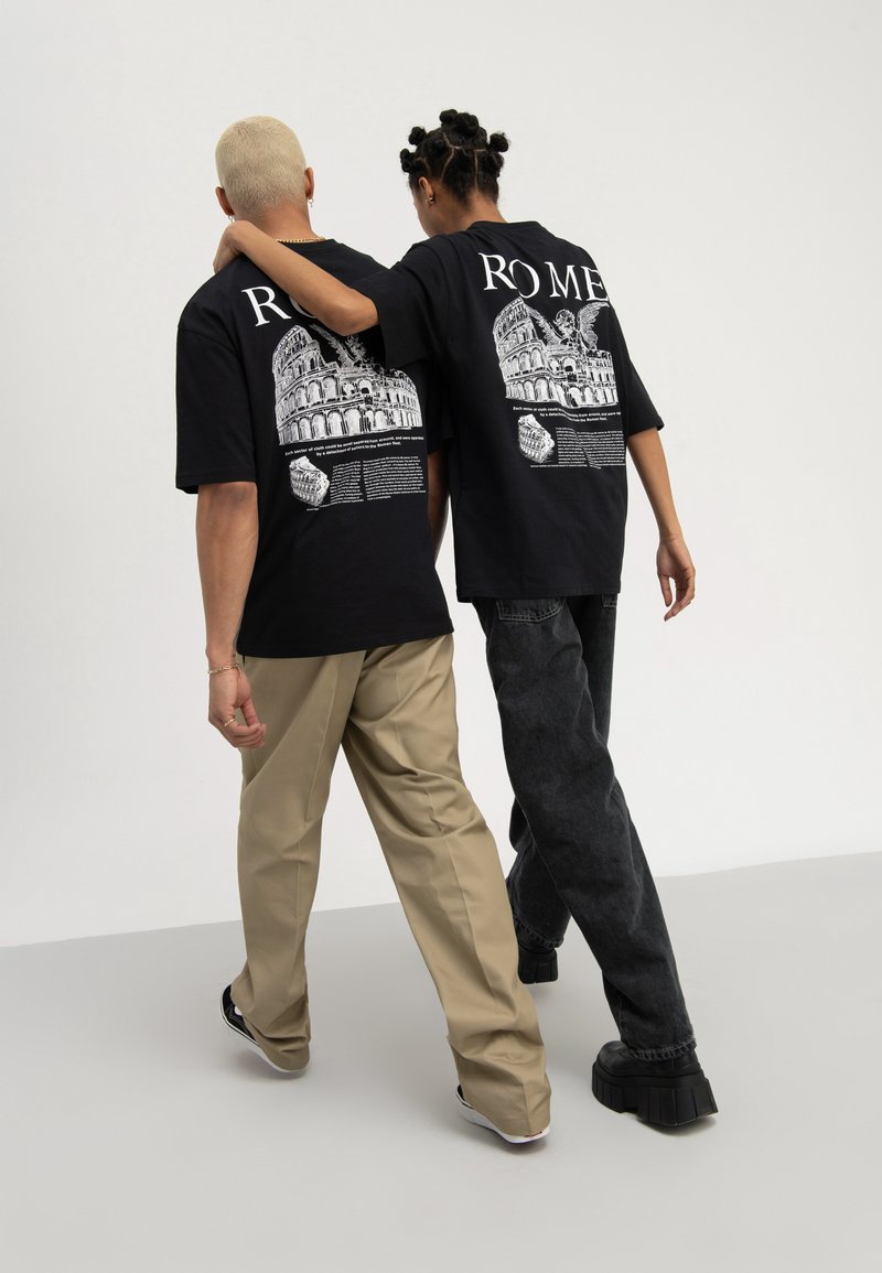 Due persone che camminano fianco a fianco con le braccia intorno alle spalle, indossando T-shirt nere con la scritta "ROME" e pantaloni casual.