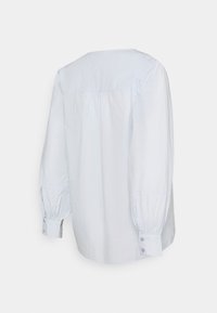 Blouse à manches longues bleu clair en coton, avec un col arrondi, des plis au dos et des boutons décoratifs sur les manches.