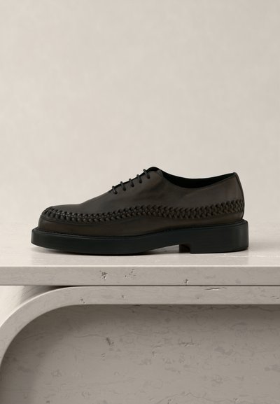 Σκούρο καφέ δερμάτινο παπούτσι με κορδόνια και πλεκτή λεπτομέρεια κατά μήκος των πλευρών, τοποθετημένο σε ανοιχτόχρωμη, υφασμένη πέτρινη επιφάνεια.