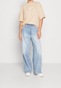 Beige bavlněné tričko s potiskem "PACIFIC COAST" v kombinaci s světle modrými kalhotami s širokými nohavicemi. Model má na sobě bílé tenisky. Jednoduché pozadí.