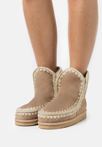 Stivaletti alla caviglia color sabbia in suede con fodera in peluche e bordi intrecciati a contrasto, caratterizzati da una punta arrotondata e suola piatta.