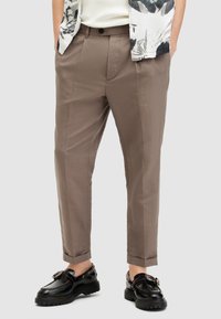 Hombre que lleva pantalones en tono topo de pierna recta con bolsillos frontales, dobladillos acampanados, combinados con mocasines negros de suela gruesa y una camisa de patrón en blanco y negro.