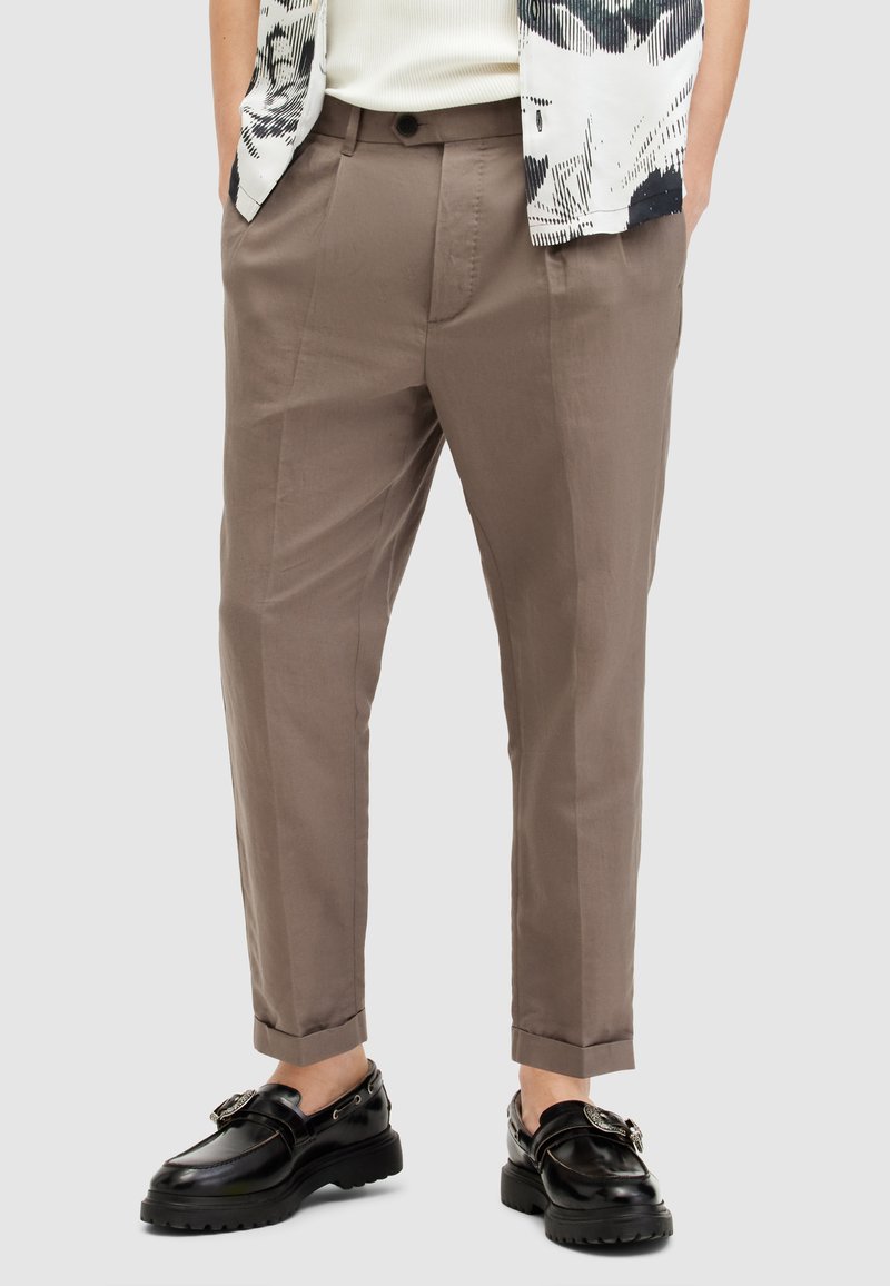Hombre que lleva pantalones en tono topo de pierna recta con bolsillos frontales, dobladillos acampanados, combinados con mocasines negros de suela gruesa y una camisa de patrón en blanco y negro.