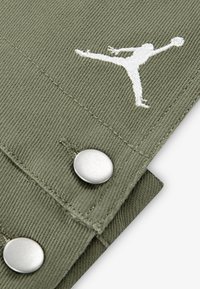 Tkanina maslinaste boje s teksturiranim završetkom, s bijelim izvezenim Jumpman logom i sjajnim metalnim kopčama.