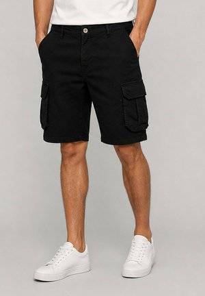 Homme portant un short cargo noir avec des poches latérales et des baskets blanches, debout les mains dans les poches devant un fond uni.