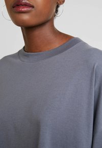 Nærbillede af en person, der bærer hoopearinger og en grå T-shirt med rund hals, hvor læber, nakke, øre og en del af skulderen vises.