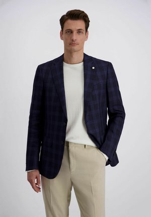 Man die een marineblauw geruit colbert draagt, een wit overhemd en beige broek, staand met één hand in de zak tegen een effen witte achtergrond.