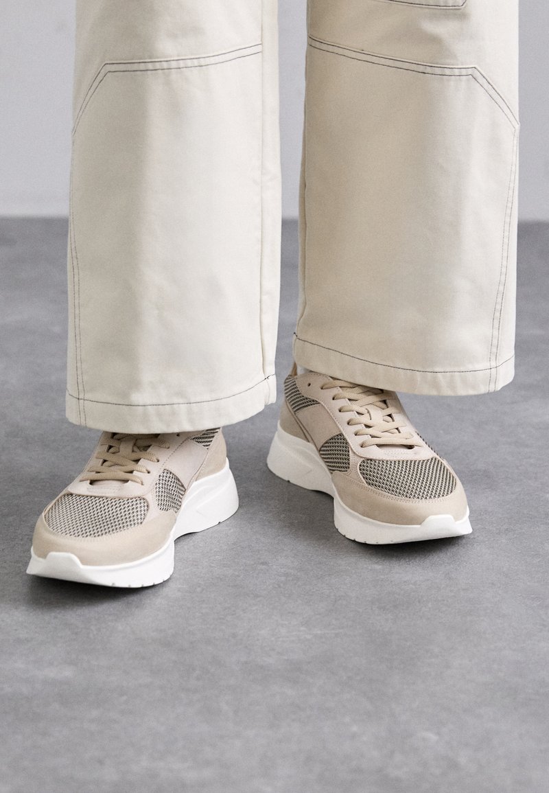 Bež športni čevlji z mrežastimi paneli in semišnimi detajli. Imajo belo debelo podplato in spredaj vezalke. Ujemajo se z kremnimi širokimi hlačami.