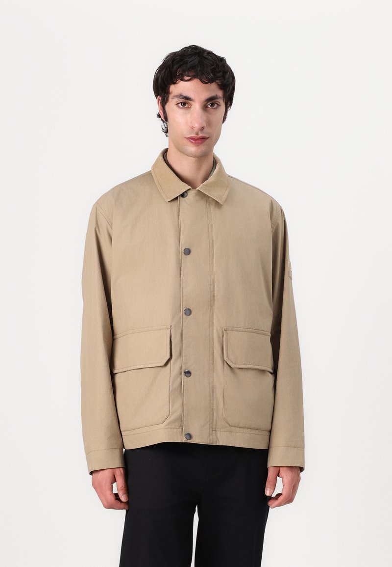 Beige canvas jacket with a collar, two large front pockets, front snap closures, and a relaxed fit. Smooth texture with minimal detailing.