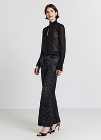 Blusa nera trasparente a maniche lunghe con fiocco al collo, abbinata a jeans neri con decorazioni in borchie d'argento sulla parte anteriore. Sandali neri con tacco alto.