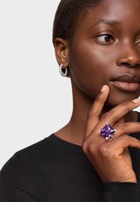 Anillo de gema púrpura con acentos plateados, que presenta una piedra cuadrada y una banda suave. Se lleva junto a un pendiente de aro con cuentas.