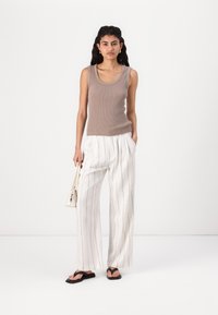 En singlet i lys brun ribbestrikket stoff, kombinert med hvite vide stripete bukser. Modellen holder en liten hvit veske. Sorte flip-flops.