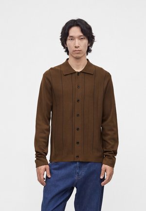 Mand med mellemlangt mørkt hår iført en brun cardigan med knapper med lodrette striber og blå jeans mod en ensfarvet baggrund.