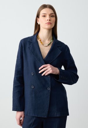 Marineblaues, doppelt geknöpftes Leinen-Sakko mit breiten Revers und Knopfakzenten, kombiniert mit einer mehrfarbigen Perlenkette. Sanfte Textur.