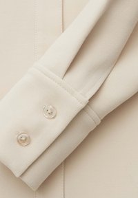 Manica di tessuto beige con una texture liscia, caratterizzata da un polsino piegato e due bottoni chiari con quattro fori ciascuno.