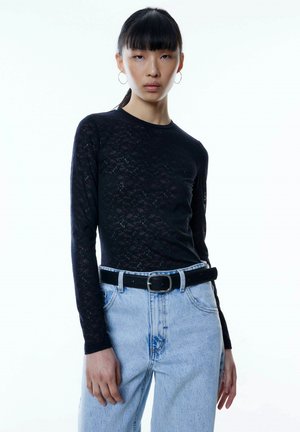 Zwarte lange mouwen top met een kanten patroon, gecombineerd met lichtblauwe high-waisted jeans en een zwarte riem met een zilveren gesp.