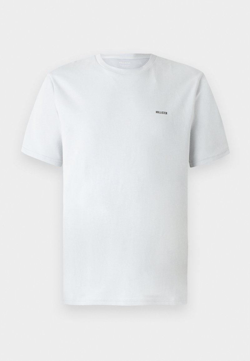 Camiseta blanca de algodón con mangas cortas, que presenta un pequeño logo negro de "Hollister" en la parte superior izquierda. Corte regular, textura suave.