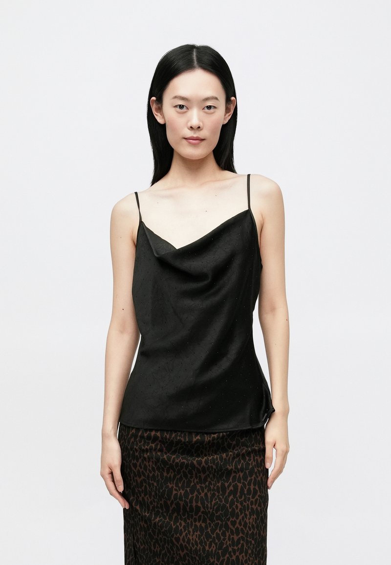 Vrouw met steil zwart haar, een zwart mouwloos shirt en een donkerbruine rok met luipaardprint, staand tegen een effen lichte achtergrond.