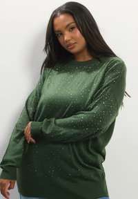 Frau mit langen dunklen Haaren, die einen dunkelgrünen Pullover mit kleinen weißen Punkten trägt, posiert vor einer schlichten hellen Wand.