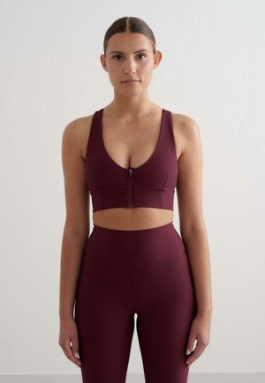 Sostenitore sportivo bordeaux a coste con scollatura a V profonda e zip frontale, abbinato a leggings a vita alta coordinati. Materiale liscio e elastico.