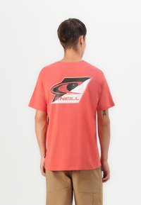 Young man seen from back wearing coral T-shirt with large black and white O'Neill logo and khaki shorts against white background.