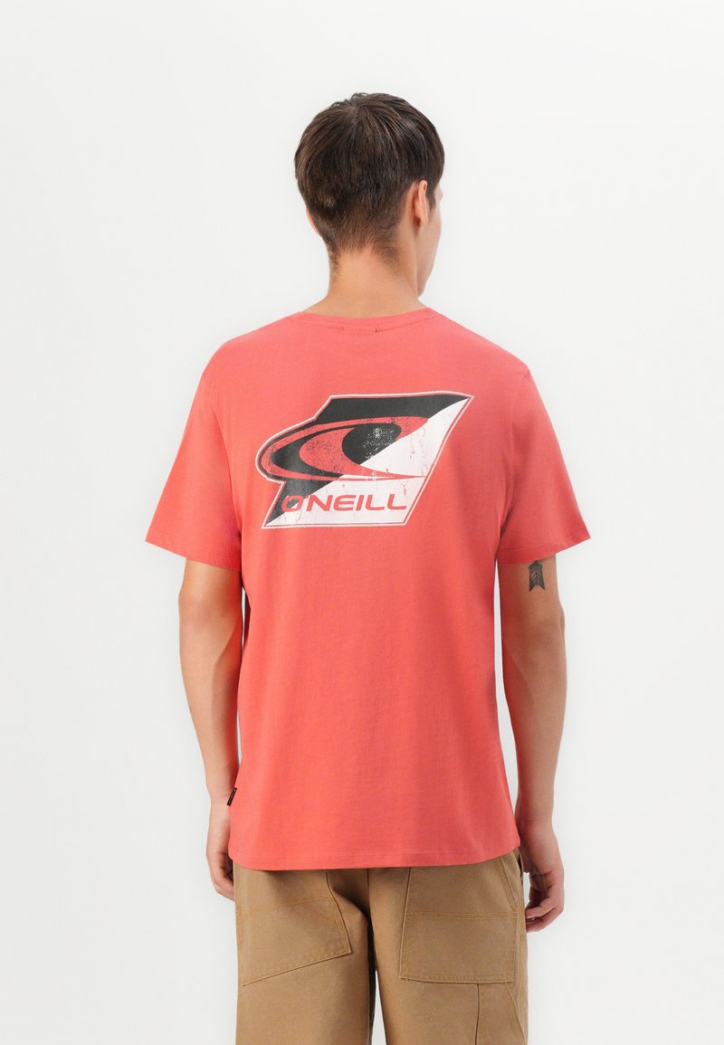 Young man seen from back wearing coral T-shirt with large black and white O'Neill logo and khaki shorts against white background.
