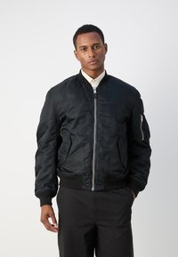 Schwarze Bomberjacke aus glattem, leichtem Material; verfügt über einen Frontreißverschluss, gerippte Bündchen und Taschen an den Seiten und dem Sleeve.
