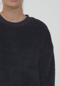 Tummansininen fleece-huppari, jossa on teksturoitu, pörröinen pinta, pyöreä kaulus ja ribbireunus. Rento muotoilu, ei näkyvää brändäystä.